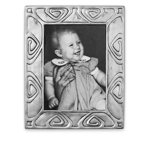 May include: A silver picture frame with an ornate design featuring a repeating swirl pattern. The frame holds a black and white photo of a baby girl smiling.