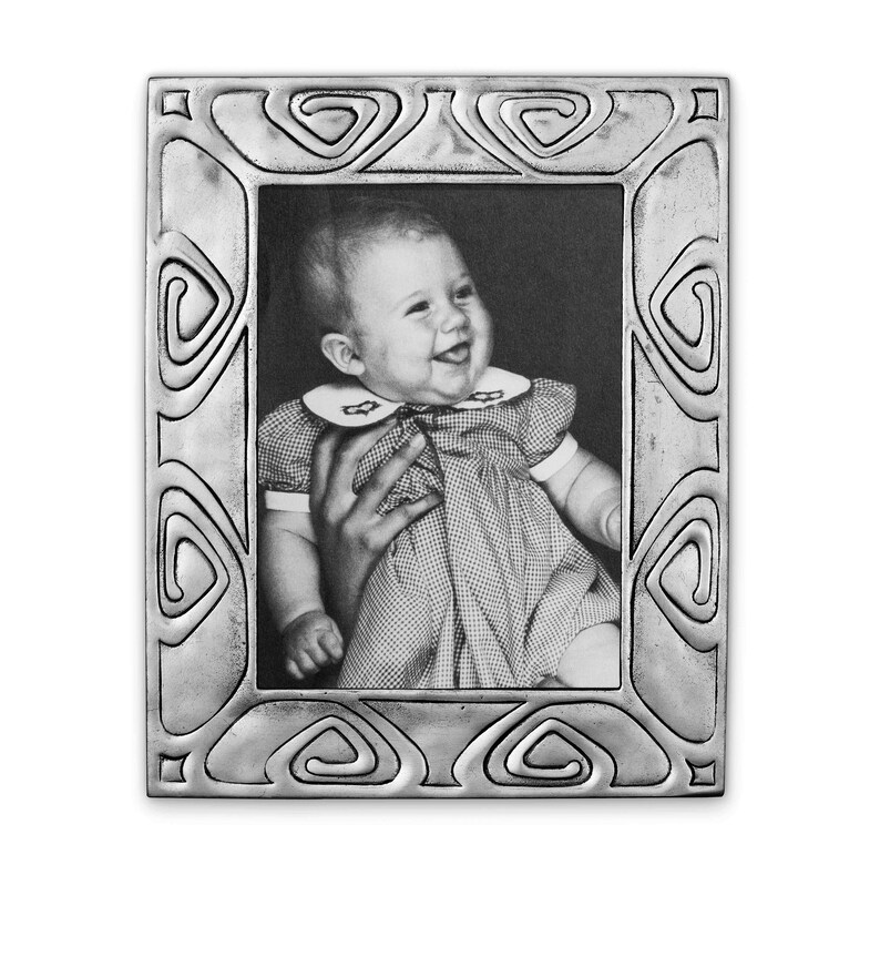 May include: A silver picture frame with an ornate design featuring a repeating swirl pattern. The frame holds a black and white photo of a baby girl smiling.
