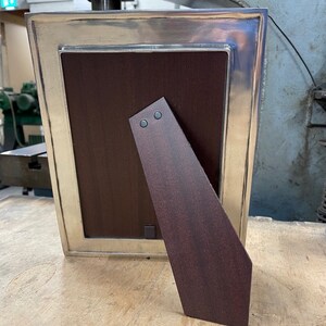 May include: A silver picture frame with a dark wood backing and a kickstand. The frame has a decorative border and a smooth finish.