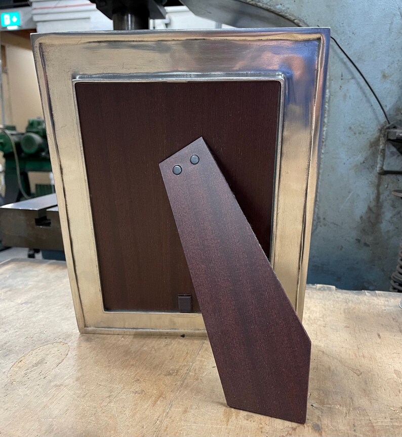 May include: A silver picture frame with a dark wood backing and a kickstand. The frame has a decorative border and a smooth finish.