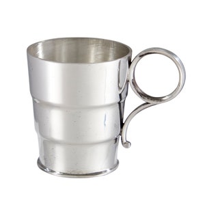 May include: A silver cup with a rounded handle and a cylindrical body. The cup has a smooth, polished finish and appears to be made of solid metal.