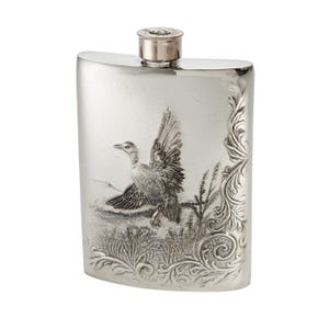 May include: A silver-toned metal flask featuring an embossed design of a duck taking flight over marshy reeds. The right side of the flask is decorated with intricate scrollwork patterns. The cap is shaped like a metallic bullet casing.