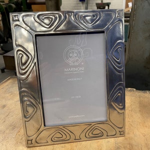 May include: A silver picture frame with an ornate design featuring a repeating pattern of circles and ovals. The frame is made in Italy by Marinoni, a company that creates objects in pewter. The frame measures 13x18 cm.