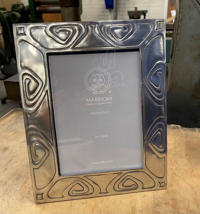 May include: A silver picture frame with an ornate design featuring a repeating pattern of circles and ovals. The frame is made in Italy by Marinoni, a company that creates objects in pewter. The frame measures 13x18 cm.