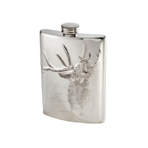 May include: A silver-colored metal flask with a curved rectangular shape and a screw-top cap. The front surface features a detailed embossed relief of a stag with large antlers, depicted in a roaring pose against a textured mountain landscape.