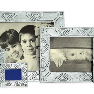 May include: Set of five silver-toned picture frames with ornate designs. The frames are arranged in a row, with the largest frame in the center and the smallest frame on the left. Each frame features a different design, but all have a similar swirling pattern. The frames are empty, but there are images of children in the background.