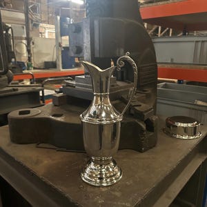 Könnte beinhalten: Eine silberne Kanne mit einem dekorativen Griff steht auf einem Metalltisch in einer Fabrikumgebung. Die Kanne hat einen runden Körper und einen ausgestellten Boden.