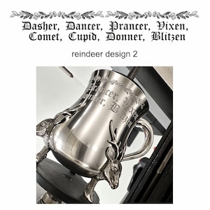 May include: A polished silver-toned metal mug featuring two sculpted reindeer heads as decorative accents at the base. The surface is engraved with the text "Prancer, Vixen, Donner, Blitzen" in a gothic font, framed by a delicate etched garland.