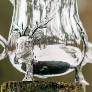 May include: A polished silver pitcher featuring a three-dimensional deer head sculpture integrated into the body. The metallic surface reflects the surroundings, and the base of the pitcher is supported by sculpted deer legs with detailed hooves.
