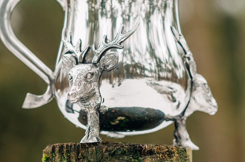May include: A polished silver pitcher featuring a three-dimensional deer head sculpture integrated into the body. The metallic surface reflects the surroundings, and the base of the pitcher is supported by sculpted deer legs with detailed hooves.