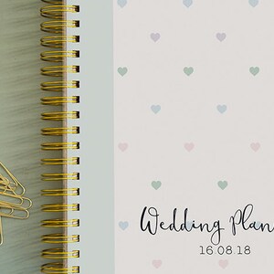 Może przedstawiać: Biały planer ślubny ze złotym spiralnym oprawieniem. Okładka ma wz&oacute;r małych pastelowych serduszek i napis "Wedding Planner" z datą "16.08.18" poniżej.