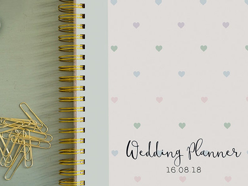 Może przedstawiać: Biały planer ślubny ze złotym spiralnym oprawieniem. Okładka ma wz&oacute;r małych pastelowych serduszek i napis "Wedding Planner" z datą "16.08.18" poniżej.