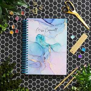 May include: A blue and pink spiral bound notebook with a marbled design and gold accents. The notebook has the text "Miss Connell" written on the front cover.