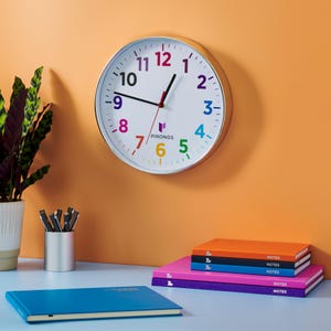 May include: A round wall clock with a white face and a silver rim. The clock has colourful numbers and black hands. The brand name "PIRONGS" is printed on the clock face. The clock is hanging on an orange wall. There are notebooks and a plant on the desk.