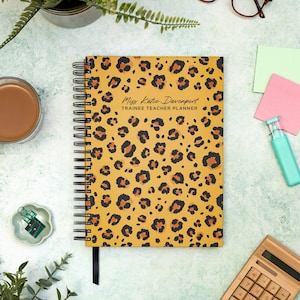 May include: A spiral-bound planner with a mustard yellow cover featuring a leopard print pattern. The text "Miss Katie Davenport TRAINEE TEACHER PLANNER" is printed on the cover. A black ribbon bookmark is attached.