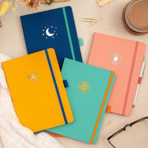 May include: Four different colored notebooks with gold foil designs. The notebooks are blue, yellow, teal, and coral. The designs are a crescent moon, a bee, a compass, and a sun.