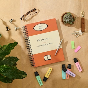 May include: A red and orange spiral bound notebook with the text "Classic Book" and "Ms Stewart Teacher Planner". The notebook is on a wooden surface with a pair of glasses, a plant, and a set of highlighters.
