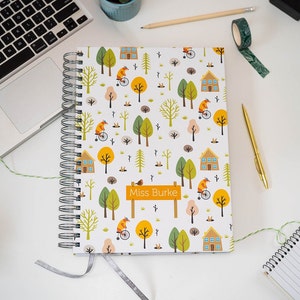 A4 or A5 wire bound Teacher planner, woodland scene of trees, animals on a penny farthing bike, houses, and rabbits peeking from a hole in the ground. Silver wire and silver lame ribbon.