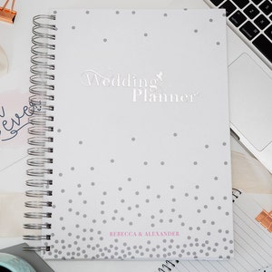 Może przedstawiać: Biały planer ślubny z szarym wzorem w kropki i tekstem "Wedding Planner" w srebrnych literach. Planer ma srebrne spiralne oprawienie, a na dole nadrukowane są imiona "Rebecca & Alexander" w kolorze r&oacute;żowym.