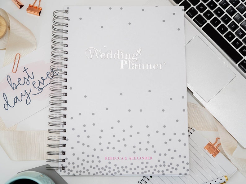 Może przedstawiać: Biały planer ślubny z szarym wzorem w kropki i tekstem "Wedding Planner" w srebrnych literach. Planer ma srebrne spiralne oprawienie, a na dole nadrukowane są imiona "Rebecca & Alexander" w kolorze r&oacute;żowym.