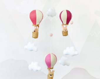 May include: A hanging mobile featuring three hot air balloons with pink and cream fabric panels. Each balloon carries a small tan plush animal in a woven basket. White felt clouds and small wooden beads are suspended on white strings.