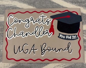 May include: A brown paper sign with a red scalloped border. The sign says "Congrats Chandler UGA Bound" in white script. A black graduation cap with a red tassel is on the sign. A white banner below the cap says "You Did It!"