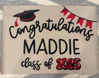 May include: A brown paper sign with black marker writing that says "Congratulations Maddie Class of 2025". There is a black graduation cap with a red band and a red string hanging from it. There are red paper flags hanging from a string above the word "Congratulations".
