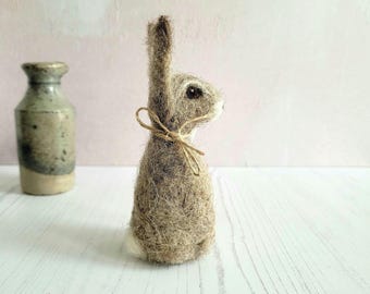 Puede incluir: Un conejo de fieltro de aguja marrón y blanco con una nariz de fieltro marrón y ojos negros. El conejo está atado con un lazo de cuerda marrón alrededor del cuello.
