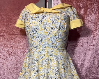May include: A vintage-style dress with a floral pattern. The dress features a yellow collar and short sleeves with yellow trim. The fabric is white with a pattern of yellow and blue leaves. The dress has a fitted bodice and a flared skirt. Bust 50 inches, waist 41 inches.