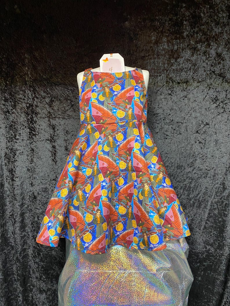 May include: A sleeveless dress on a mannequin with a colorful pattern. The dress features a repeating design of figures with wings, set against a blue, red, and yellow background. The dress has a flared skirt.