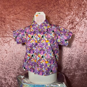 May include: Purple short-sleeved button-down shirt with a cartoon dragon print. The shirt has a tag that says "12-18".