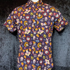May include: Short-sleeved button-down shirt with a dark purple background and a colorful pattern of skulls, musical notes, and floral designs. The shirt has a chest pocket and is displayed on a mannequin.