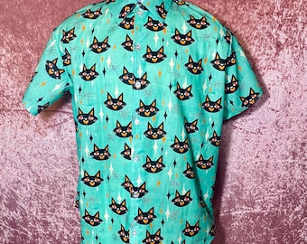 May include: A turquoise blue short-sleeved button-down shirt with a black cat face pattern and gold star accents. The shirt has a collar and a button closure. The size tag says "XL".