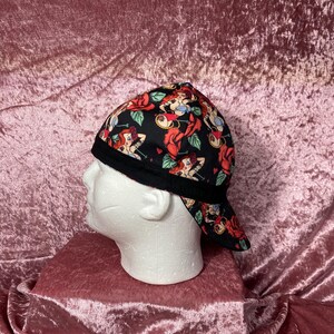 May include: Black cap with a pattern of red roses, green leaves, and pin-up girl illustrations. The cap has a black brim and is displayed on a white mannequin head. The background is a pink, textured fabric.