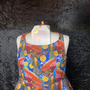 May include: A sleeveless dress with a vibrant print featuring a repeating pattern of a person with a futuristic vehicle against a blue and starry background. The dress has a scoop neckline and a flared skirt. The dress is displayed on a mannequin.