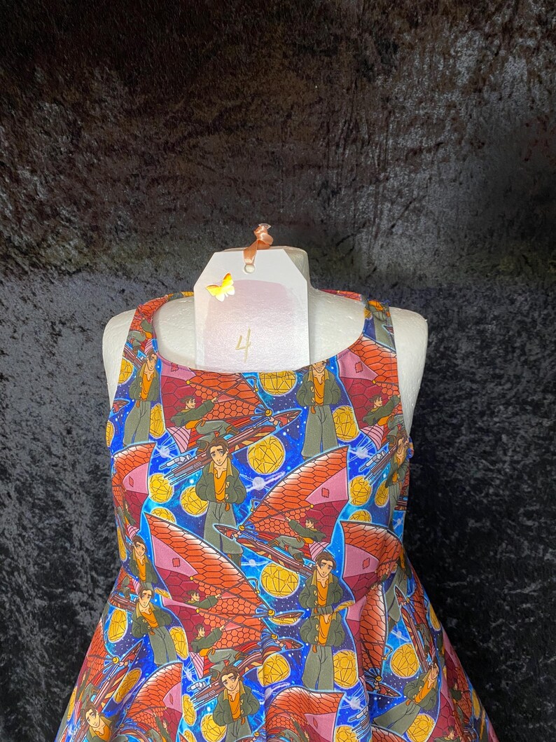 May include: A sleeveless dress with a vibrant print featuring a repeating pattern of a person with a futuristic vehicle against a blue and starry background. The dress has a scoop neckline and a flared skirt. The dress is displayed on a mannequin.
