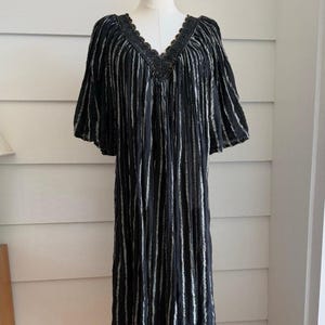 Könnte beinhalten: Ein schwarzes, wadenlanges Kleid mit vertikalem weißem Streifenmuster, V-Ausschnitt mit dunkler, gebogter Spitzenborte und locker sitzenden, ellbogenlangen Ärmeln. Das Kleidungsstück hat eine geraffte, fließende Silhouette aus strukturiertem Knitterstoff.