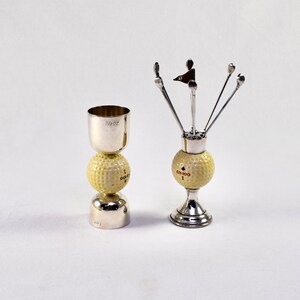 Könnte beinhalten: Zwei silberne Golfball-Schnapsgläser. Ein Schnapsglas ist ein Standard-Schnapsglas mit einem Golfball obenauf. Das andere Schnapsglas hat einen Golfball-Sockel mit fünf silbernen Golfschläger-Stiften, die aus der Oberseite herausragen. Der Golfball auf dem Sockel trägt den Text "GX-100 1".