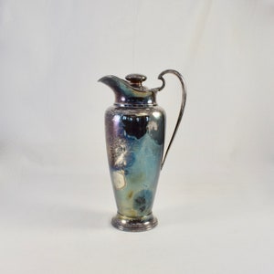 Puede incluir: Una jarra de plata con cuerpo redondeado y tapa. La jarra tiene asa y base ligeramente acampanada.