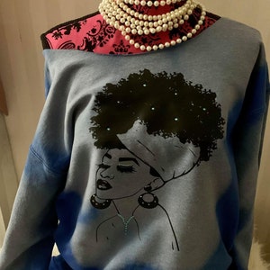 May include: A blue tie-dye sweatshirt with a black and white graphic of a woman with an afro and a headscarf. The woman is wearing earrings and a necklace with turquoise accents.