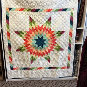 May include: A square quilt featuring a central eight-pointed star pattern in shades of red, orange, green, and teal. The star is surrounded by a white background with a quilted wave texture and a multi-colored striped border in a diagonal pattern.