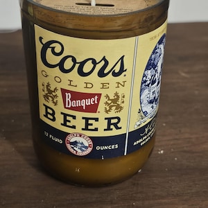 May include: A scented candle in a glass jar with a label featuring the Coors Golden Banquet Beer logo. The label includes text such as "12 Fluid Ounces" and "Adolph Coors Golden, Colorado."