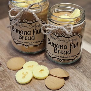 May include: Two glass jars of Banana Nut Bread scented soy blend candles with banana slices on top. The jars are tied with twine and have labels that read "Rubber Charm Creations". Several cookies and banana slices are on a wooden board.