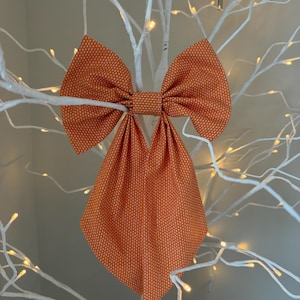 May include: A large orange fabric bow featuring a repeating pattern of small white vertical dashes. The bow has two wide, pointed tails hanging downward and a central knot, constructed from a textured material with a consistent orange and white design.
