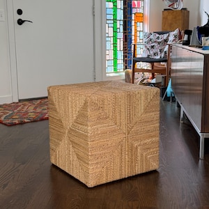 May include: A square ottoman constructed from tightly coiled natural tan seagrass fibers. The exterior surface features a geometric pattern created by the directional orientation of the woven material, forming a series of radiating square shapes.