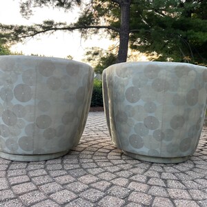 Op de afbeelding: Twee lichtgrijze, afgeronde plantenbakken met een cirkelvormig patroon. De plantenbakken zijn gemaakt van een textuurrijk materiaal en staan op een cirkelvormige basis. De achtergrond omvat bomen en een stenen terras.