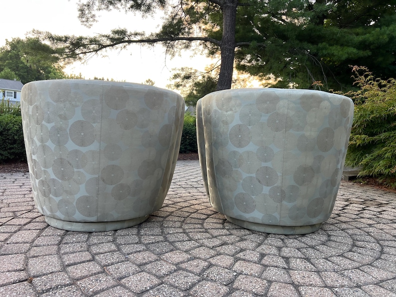 Op de afbeelding: Twee lichtgrijze, afgeronde plantenbakken met een cirkelvormig patroon. De plantenbakken zijn gemaakt van een textuurrijk materiaal en staan op een cirkelvormige basis. De achtergrond omvat bomen en een stenen terras.