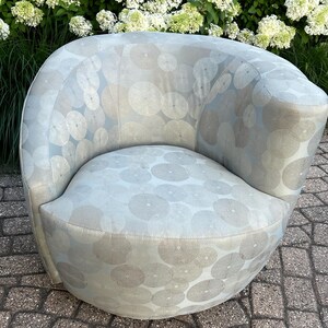 Op de afbeelding: Een lichtblauwe, afgeronde fauteuil met een cirkelvormig patroon in een lichtere tint. De stoel heeft een gebogen rugleuning en een afgeronde zitting. De stof heeft een subtiele glans. De stoel staat buiten, met groen op de achtergrond.