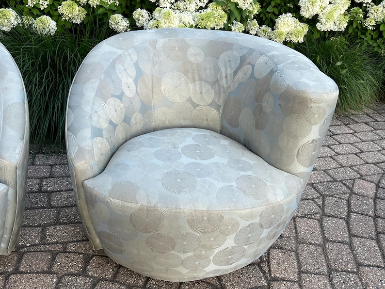 Op de afbeelding: Een lichtblauwe, afgeronde fauteuil met een cirkelvormig patroon in een lichtere tint. De stoel heeft een gebogen rugleuning en een afgeronde zitting. De stof heeft een subtiele glans. De stoel staat buiten, met groen op de achtergrond.