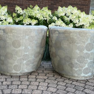 Op de afbeelding: Twee tonvormige fauteuils met een lichtgrijze stof met cirkelpatroon. De stoelen staan op een stenen terras met op de achtergrond witte hortensiastruiken en een bakstenen muur.